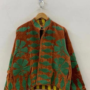 May include: A brown velvet jacket with green embroidered floral and leaf designs. The jacket has a mandarin collar and a yellow and brown striped lining. The jacket is hanging on a wooden hanger.