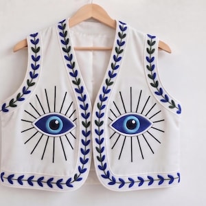 May include: A white sleeveless waistcoat featuring two large embroidered blue and black eye motifs on the front panels. The garment is detailed with a continuous blue and dark green leaf-patterned border along the neckline, armholes, and hemline.