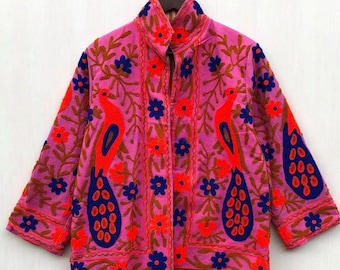Puede incluir: Chaqueta rosa con cuello alto, adornada con pájaros naranjas bordados, flores azules y tallos marrones. La chaqueta está colgada de una percha de madera, mostrando su intrincado diseño floral y de aves.