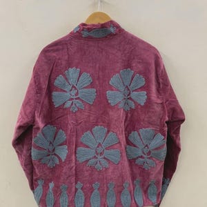 May include: A magenta jacket with a high collar and long sleeves, adorned with large, embroidered blue floral designs. The jacket is hanging on a wooden hanger against a neutral background, showcasing its detailed craftsmanship and unique style.