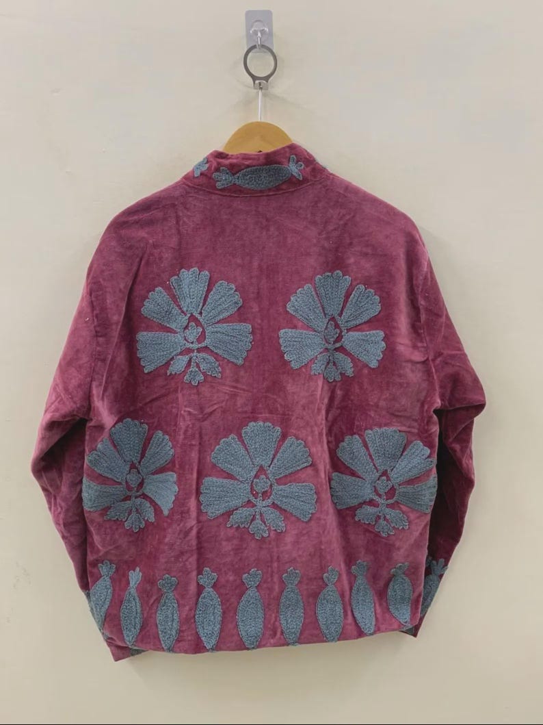 May include: A magenta jacket with a high collar and long sleeves, adorned with large, embroidered blue floral designs. The jacket is hanging on a wooden hanger against a neutral background, showcasing its detailed craftsmanship and unique style.