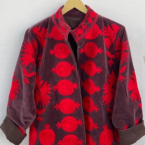Puede incluir: Chaqueta de terciopelo color ciruela con bordados florales rojos. La chaqueta presenta un cuello mandar&iacute;n, mangas tres cuartos y pu&ntilde;os marrones. El bordado rojo incluye flores estilizadas y formas circulares, creando un contraste con la tela oscura.