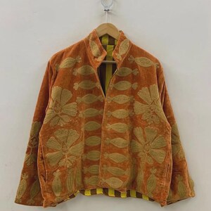 May include: An orange velvet jacket with gold floral embroidery. The jacket has a high collar and long sleeves. The interior lining is yellow and brown striped. The jacket is hanging on a wooden hanger.