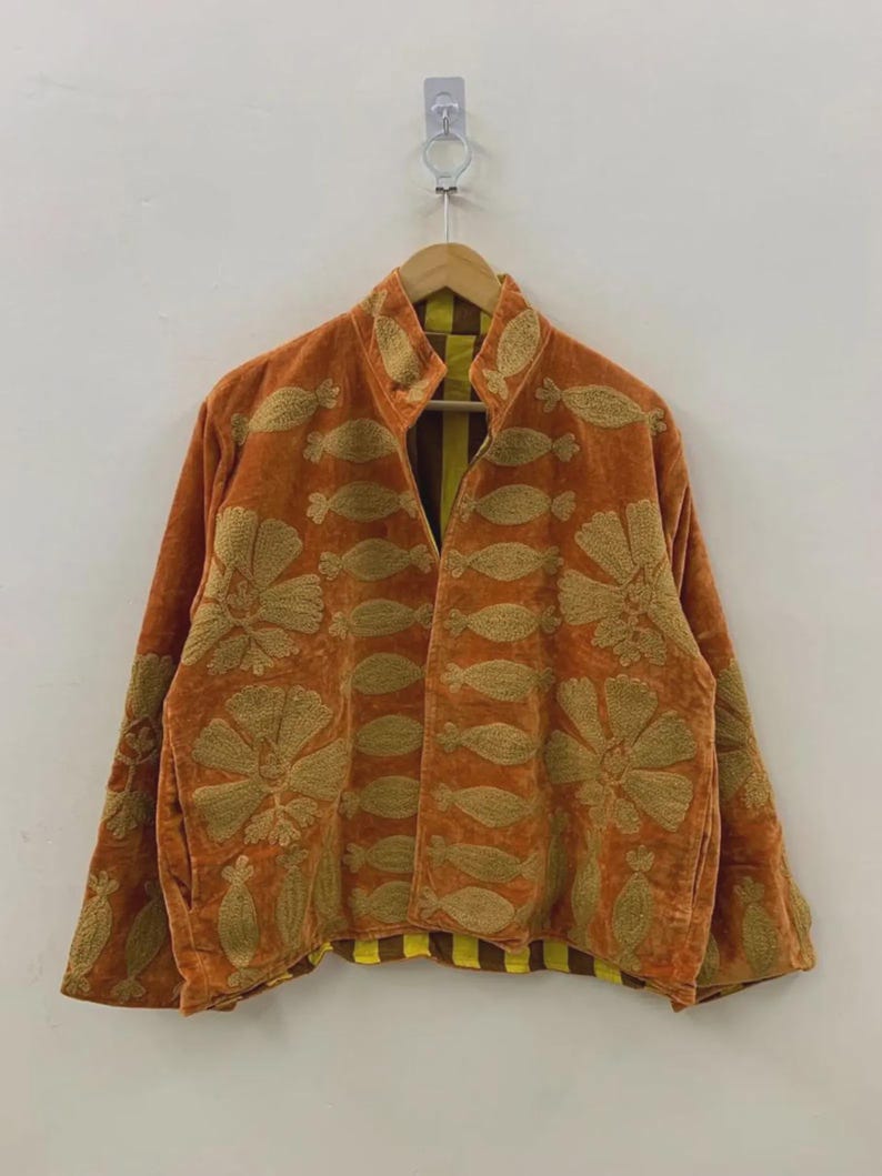 May include: An orange velvet jacket with gold floral embroidery. The jacket has a high collar and long sleeves. The interior lining is yellow and brown striped. The jacket is hanging on a wooden hanger.