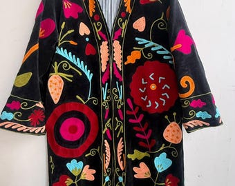 May include: A black long-sleeve coat featuring vibrant, multi-colored floral and botanical embroidery throughout. The garment has a V-neckline, a straight silhouette, and a blue and white pinstriped lining visible at the collar and interior edges.