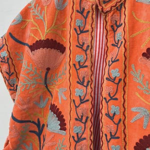 May include: An orange velvet jacket with floral embroidery in shades of blue, yellow, and burgundy. The jacket features a collared neckline and a striped red and white lining. The embroidery is detailed and covers the front and sleeves.