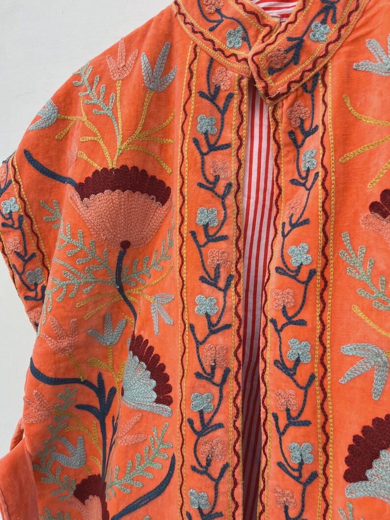May include: An orange velvet jacket with floral embroidery in shades of blue, yellow, and burgundy. The jacket features a collared neckline and a striped red and white lining. The embroidery is detailed and covers the front and sleeves.