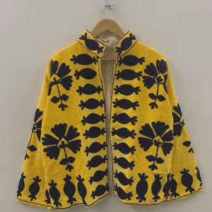 May include: A yellow jacket with a floral design in navy blue. The jacket has a mandarin collar and is open in the front. The floral pattern is repeated throughout the jacket. The jacket is hanging on a wooden hanger.