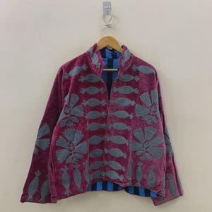 May include: A burgundy velvet jacket with gray embroidered floral and abstract designs. The jacket has a high collar and long sleeves. The interior lining is blue and black striped. The jacket is hanging on a wooden hanger.