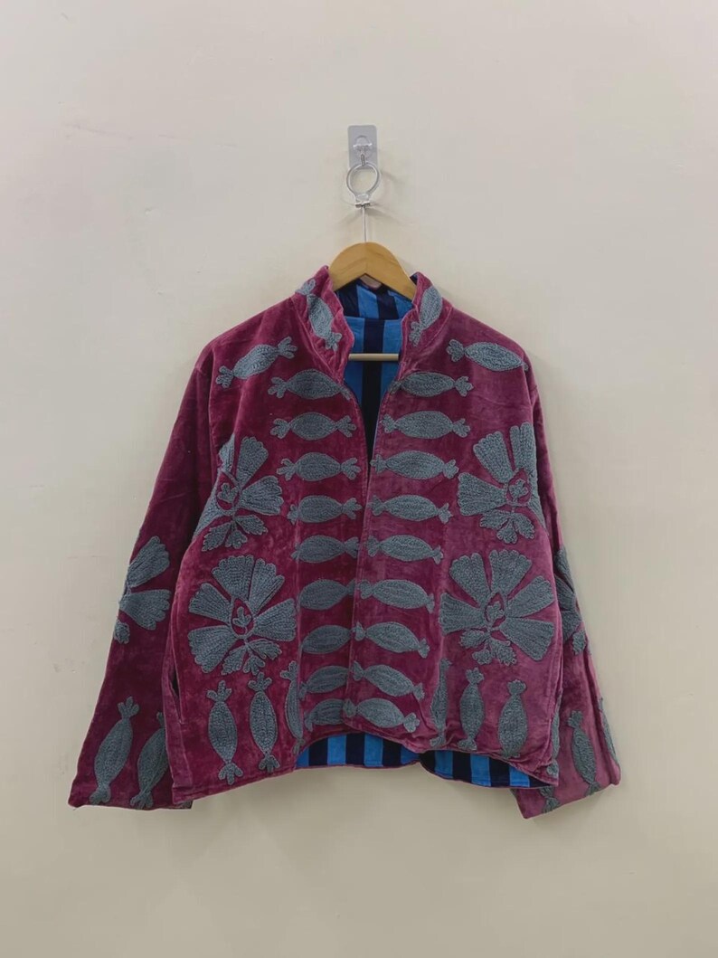May include: A burgundy velvet jacket with gray embroidered floral and abstract designs. The jacket has a high collar and long sleeves. The interior lining is blue and black striped. The jacket is hanging on a wooden hanger.