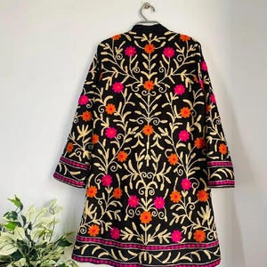 May include: Black embroidered jacket with a floral pattern in orange and pink. The jacket has long sleeves and a round collar. The embroidery uses gold thread, and it hangs on a silver hanger. The jacket is a knee-length coat.