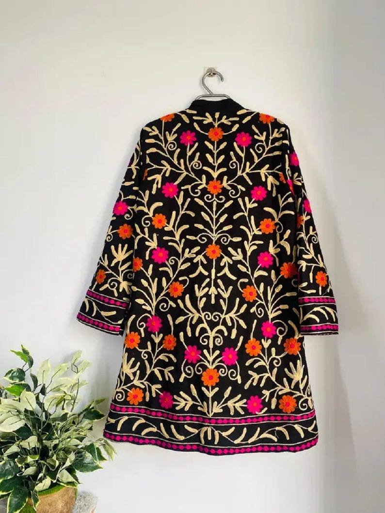 May include: Black embroidered jacket with a floral pattern in orange and pink. The jacket has long sleeves and a round collar. The embroidery uses gold thread, and it hangs on a silver hanger. The jacket is a knee-length coat.