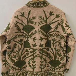 May include: Beige jacket with intricate green floral embroidery. The jacket features a high collar and long sleeves. The embroidery design includes stylized flowers and leaves, creating a detailed pattern across the back and sleeves.