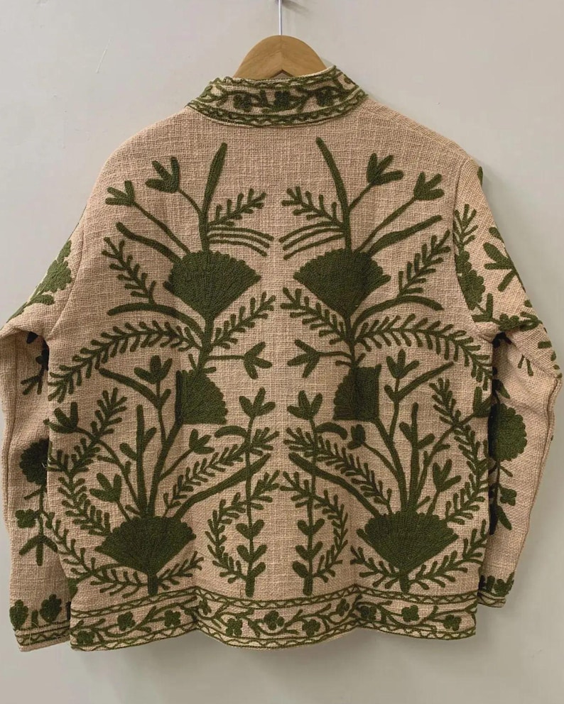 May include: Beige jacket with intricate green floral embroidery. The jacket features a high collar and long sleeves. The embroidery design includes stylized flowers and leaves, creating a detailed pattern across the back and sleeves.