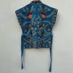 以下が含まれることがあります： フローラルデザインの、ターコイズブルーの刺繍入りベスト。ベストはラップアラウンドベルト付きで、オレンジ、ピンク、グリーンの花の刺繍が施されています。木製のハンガーにかかっています。
