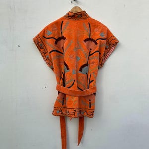 May include: An orange embroidered robe with a matching belt and tie closure. The robe features floral embroidery in shades of blue, brown, and green. The robe is hanging on a wooden hanger against a white wall.