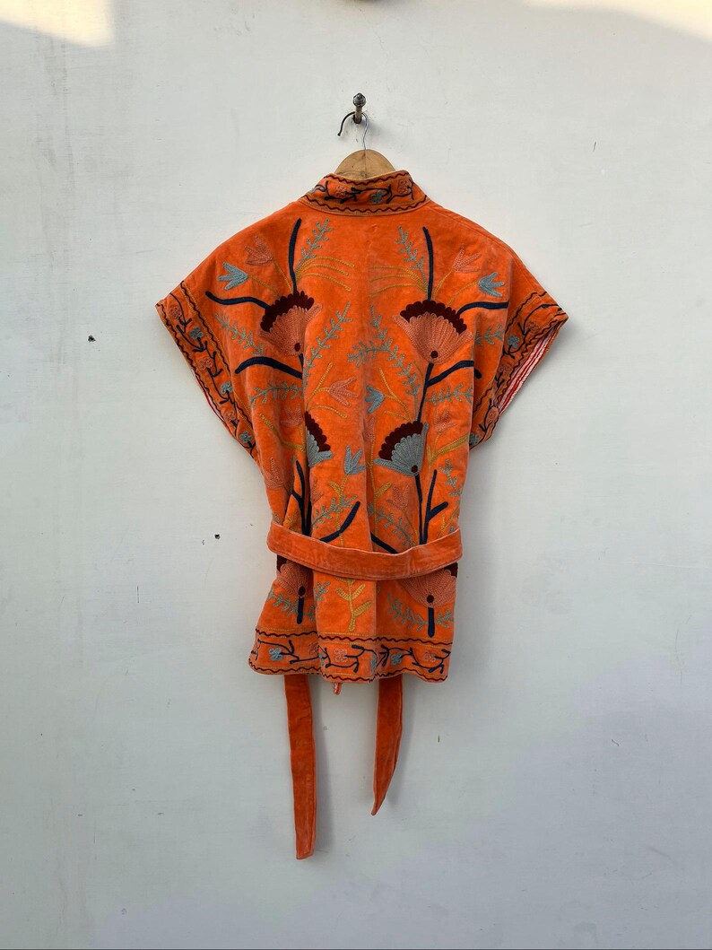 May include: An orange embroidered robe with a matching belt and tie closure. The robe features floral embroidery in shades of blue, brown, and green. The robe is hanging on a wooden hanger against a white wall.