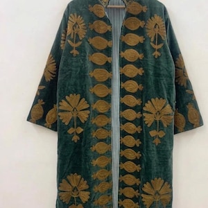 Pode incluir: Um casaco comprido e aberto em veludo verde-azulado escuro. O casaco apresenta intrincados desenhos florais e ovais bordados em dourado. O forro interior é um padrão listrado de branco e azul claro. O casaco está pendurado num cabide de madeira.