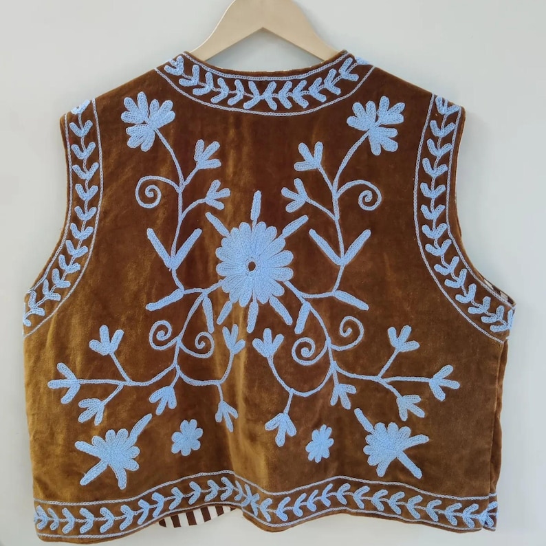 May include: Brown velvet vest with light blue floral embroidery. The sleeveless vest features a central flower design and decorative border. The embroidery is detailed and covers the front and back of the vest.