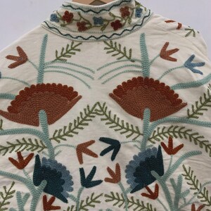 Puede incluir: Chaqueta color crema con intrincados bordados florales en tonos marr&oacute;n, azul y verde. La chaqueta presenta un cuello alto y un dise&ntilde;o sim&eacute;trico de flores y hojas estilizadas, creando un patr&oacute;n detallado y art&iacute;stico.