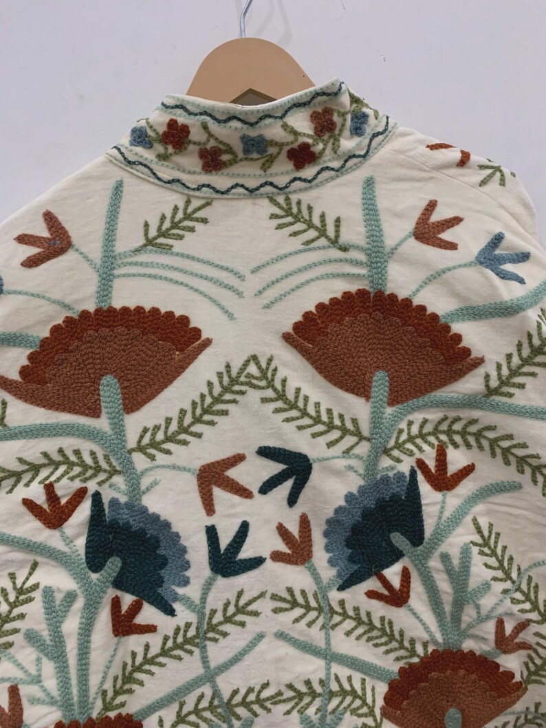 Puede incluir: Chaqueta color crema con intrincados bordados florales en tonos marr&oacute;n, azul y verde. La chaqueta presenta un cuello alto y un dise&ntilde;o sim&eacute;trico de flores y hojas estilizadas, creando un patr&oacute;n detallado y art&iacute;stico.