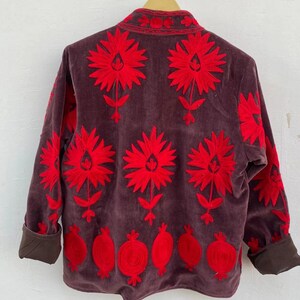 Puede incluir: Chaqueta de pana marr&oacute;n con vibrante bordado floral rojo. La chaqueta presenta grandes flores estilizadas y dise&ntilde;os de granadas. Los pu&ntilde;os y el cuello son de un marr&oacute;n m&aacute;s oscuro. La chaqueta est&aacute; colgada en una percha de madera.