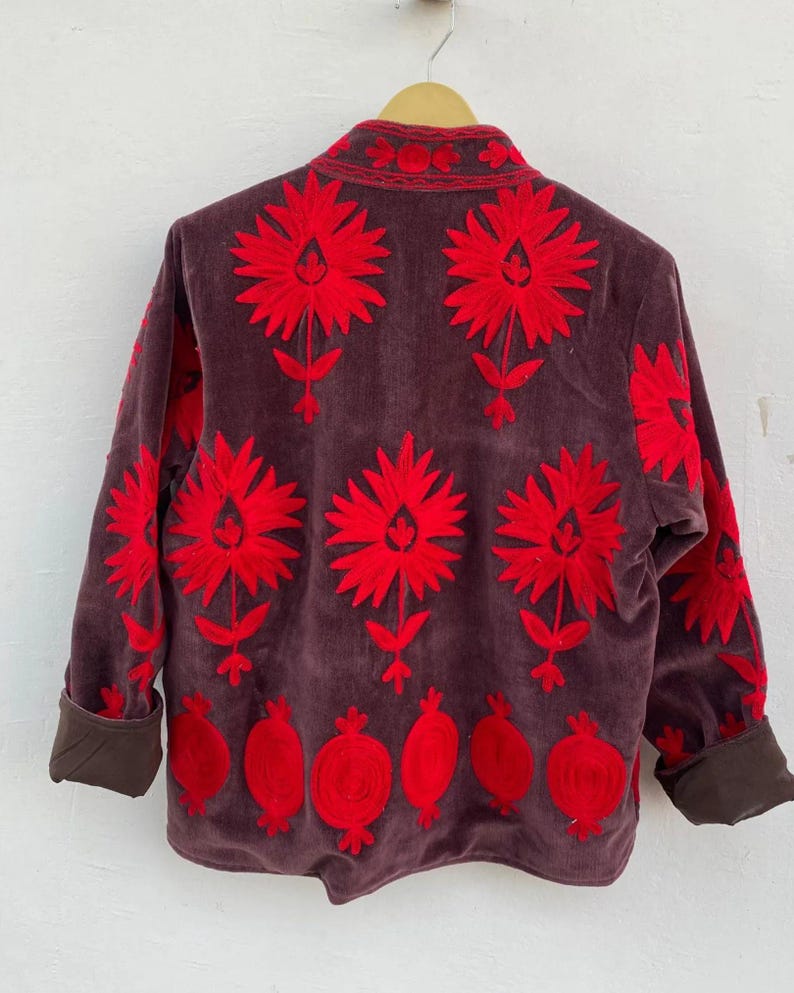 Puede incluir: Chaqueta de pana marr&oacute;n con vibrante bordado floral rojo. La chaqueta presenta grandes flores estilizadas y dise&ntilde;os de granadas. Los pu&ntilde;os y el cuello son de un marr&oacute;n m&aacute;s oscuro. La chaqueta est&aacute; colgada en una percha de madera.