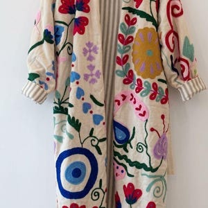 May include: Cream-colored embroidered coat with colorful floral and eye designs. The long coat features red, blue, yellow, and purple embroidered flowers and leaves. The interior lining has vertical stripes, and the sleeves have striped cuffs. The coat is hanging on a wooden hanger.