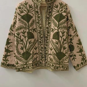 May include: A beige jacket with a stand-up collar and long sleeves. The jacket features an intricate embroidered floral design in shades of green. The interior lining has a striped pattern. The jacket is hanging on a wooden hanger.