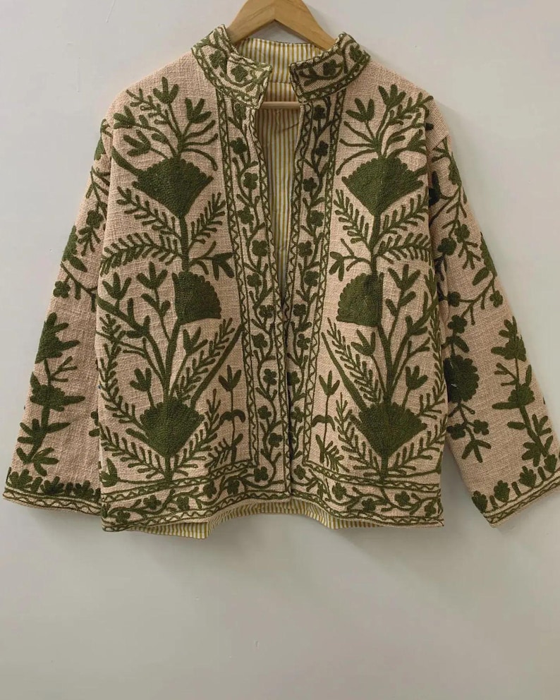 May include: A beige jacket with a stand-up collar and long sleeves. The jacket features an intricate embroidered floral design in shades of green. The interior lining has a striped pattern. The jacket is hanging on a wooden hanger.