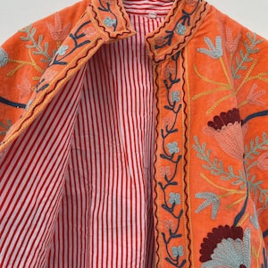 May include: An orange jacket with floral embroidery in shades of blue, yellow, and maroon. The jacket's interior features a red and white striped lining. The jacket is hanging on a wooden hanger.