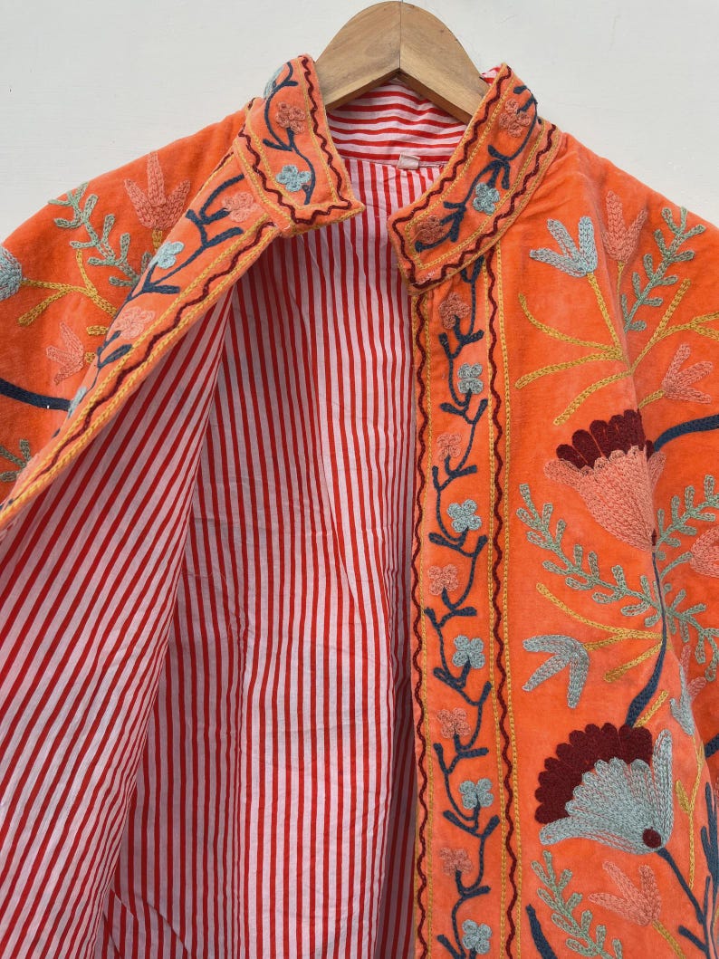 May include: An orange jacket with floral embroidery in shades of blue, yellow, and maroon. The jacket's interior features a red and white striped lining. The jacket is hanging on a wooden hanger.