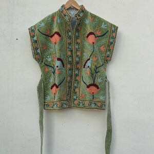 May include: A green velvet vest with floral embroidery in peach, blue, and green. The vest has a mandarin collar and a striped lining. It features a tie closure and is displayed on a wooden hanger.
