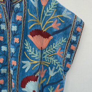 以下が含まれることがあります： ターコイズブルーの刺繍入りジャケットのクローズアップ。ジャケットは、コーラル、ライトブルー、イエローの色合いで複雑な花の刺繍が施されています。刺繍はさまざまなステッチで詳細に施されており、テクスチャード加工で視覚的に魅力的なデザインになっています。ジャケットはマンダリンカラーです。