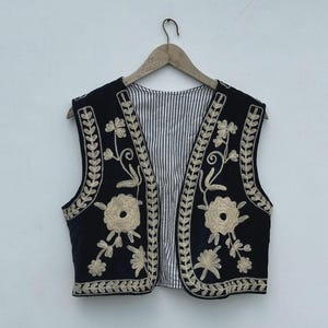 Pode incluir: Um colete preto com bordados florais creme e padrões de folhas. O colete tem um forro listrado e é exibido em um cabide de madeira. Os detalhes do bordado são visíveis na frente e nas bordas.