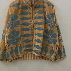 May include: A mustard-colored jacket with blue floral embroidery. The jacket has a collared neckline and a striped lining. The embroidery features a repeating pattern of stylized flowers and leaf shapes. The jacket is hanging on a wooden hanger.