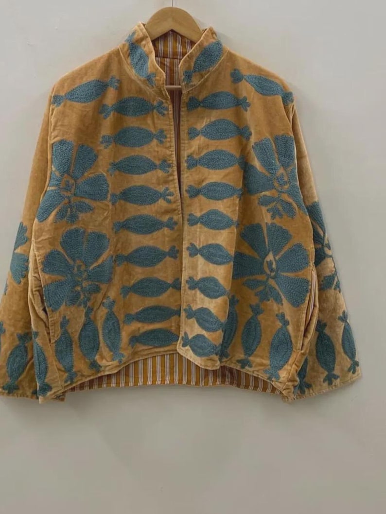 May include: A mustard-colored jacket with blue floral embroidery. The jacket has a collared neckline and a striped lining. The embroidery features a repeating pattern of stylized flowers and leaf shapes. The jacket is hanging on a wooden hanger.