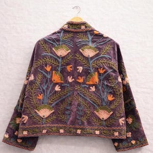 May include: A purple velvet jacket with intricate floral embroidery in shades of orange, green, and pink. The jacket is hanging on a wooden hanger, showcasing the detailed design on the back and sleeves. The embroidery features stylized flowers and leaves.