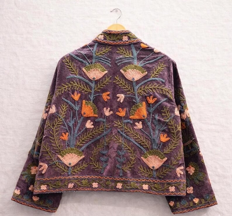 May include: A purple velvet jacket with intricate floral embroidery in shades of orange, green, and pink. The jacket is hanging on a wooden hanger, showcasing the detailed design on the back and sleeves. The embroidery features stylized flowers and leaves.