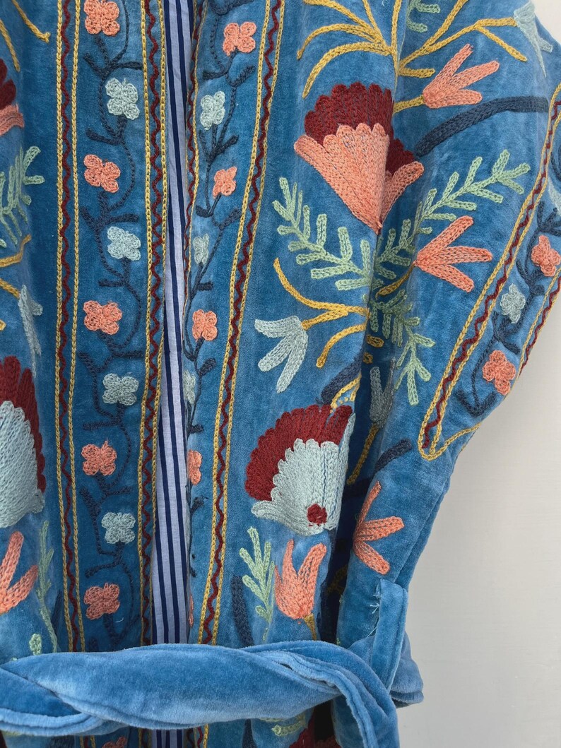 以下が含まれることがあります： 赤、桃色、黄色、緑の色合いの花柄が刺繍された青いローブのクローズアップ。 ローブにはお揃いの青いベルトが付いています。 刺繍は細かく、ローブの表面を覆っています。