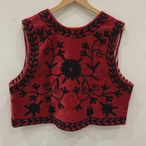 May include: A red sleeveless vest with black embroidery of floral designs. The vest has a rounded neckline and a scalloped hem.