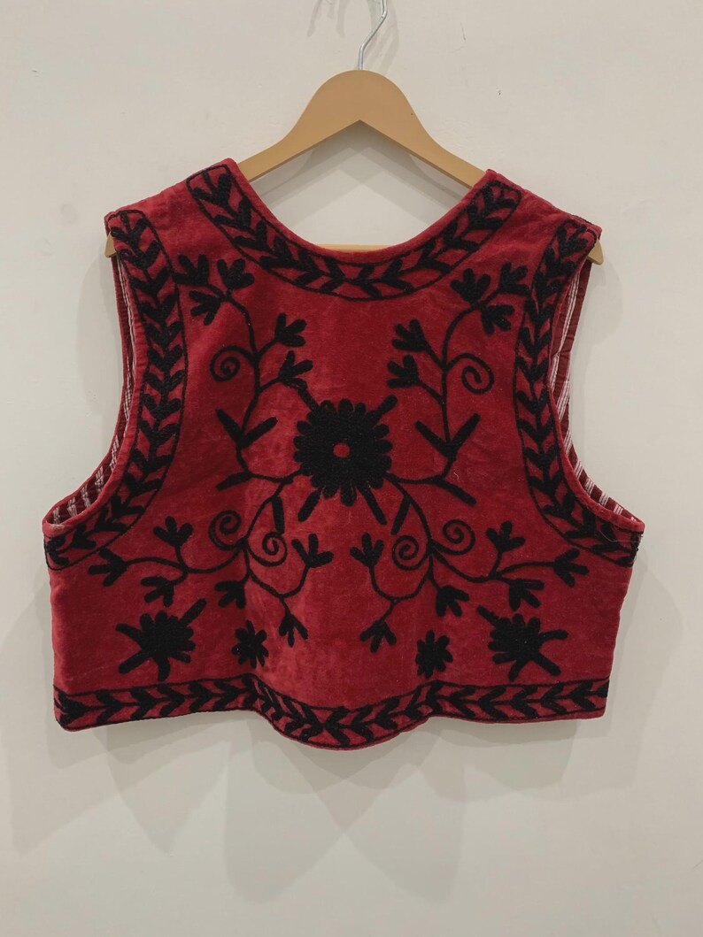 May include: A red sleeveless vest with black embroidery of floral designs. The vest has a rounded neckline and a scalloped hem.