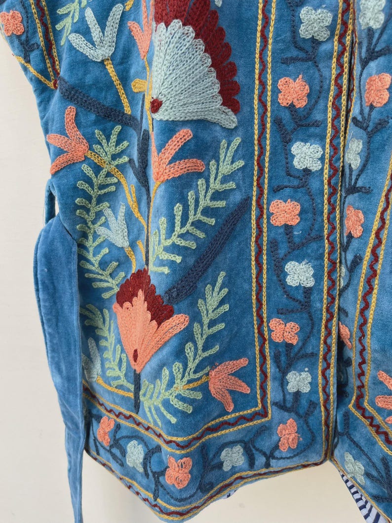 以下が含まれることがあります： 青い刺繍入りベストのクローズアップ。オレンジ、赤、緑の色合いのカラフルな花の刺繍が施されています。縁は金色の糸でトリミングされ、ベストは紐で留められています。