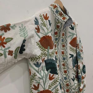 Puede incluir: Chaqueta blanca con bordados florales coloridos. La chaqueta tiene cuello y mangas largas. El bordado incluye flores y hojas naranjas, verdes, azules y marrones. La chaqueta est&aacute; colgada de una percha de madera.