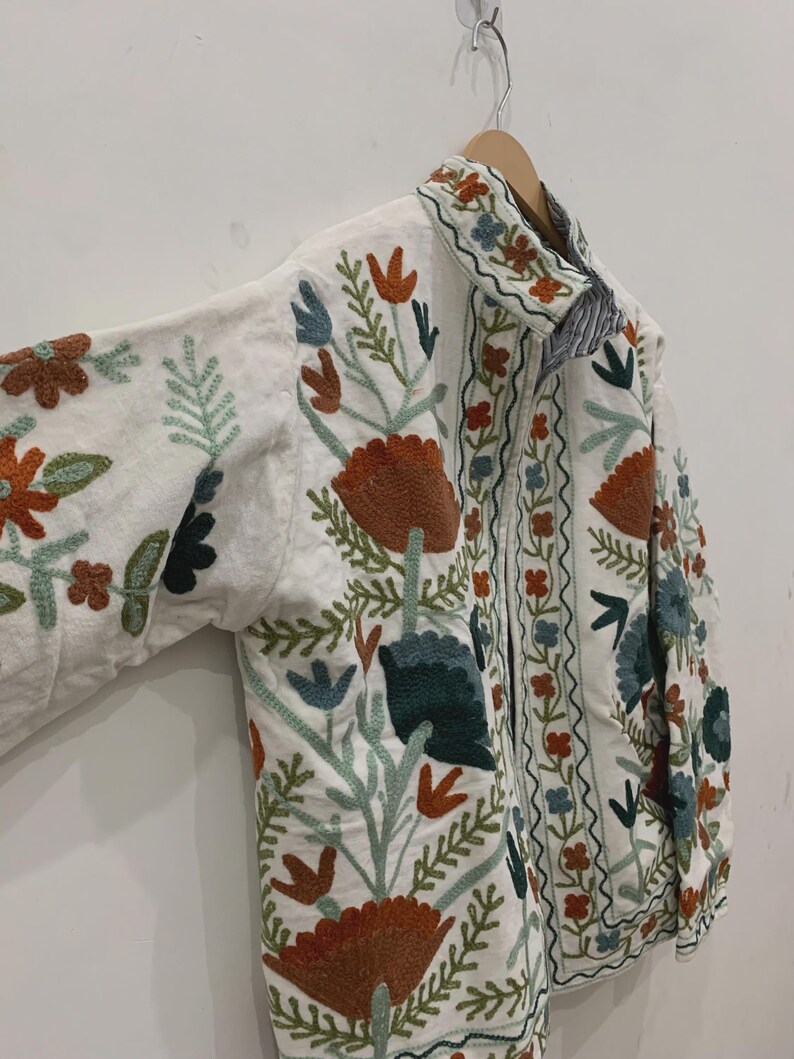 Puede incluir: Chaqueta blanca con bordados florales coloridos. La chaqueta tiene cuello y mangas largas. El bordado incluye flores y hojas naranjas, verdes, azules y marrones. La chaqueta est&aacute; colgada de una percha de madera.