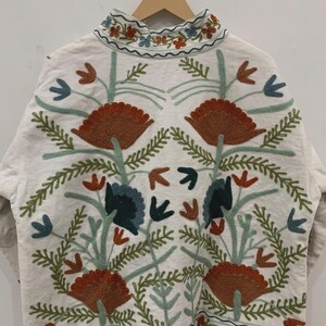 Puede incluir: Chaqueta blanca con intrincados bordados florales en tonos naranja, azul y verde. La chaqueta presenta un cuello alto y patrones detallados, mostrando un estilo bohemio o de arte popular. El bordado incluye flores y hojas estilizadas.