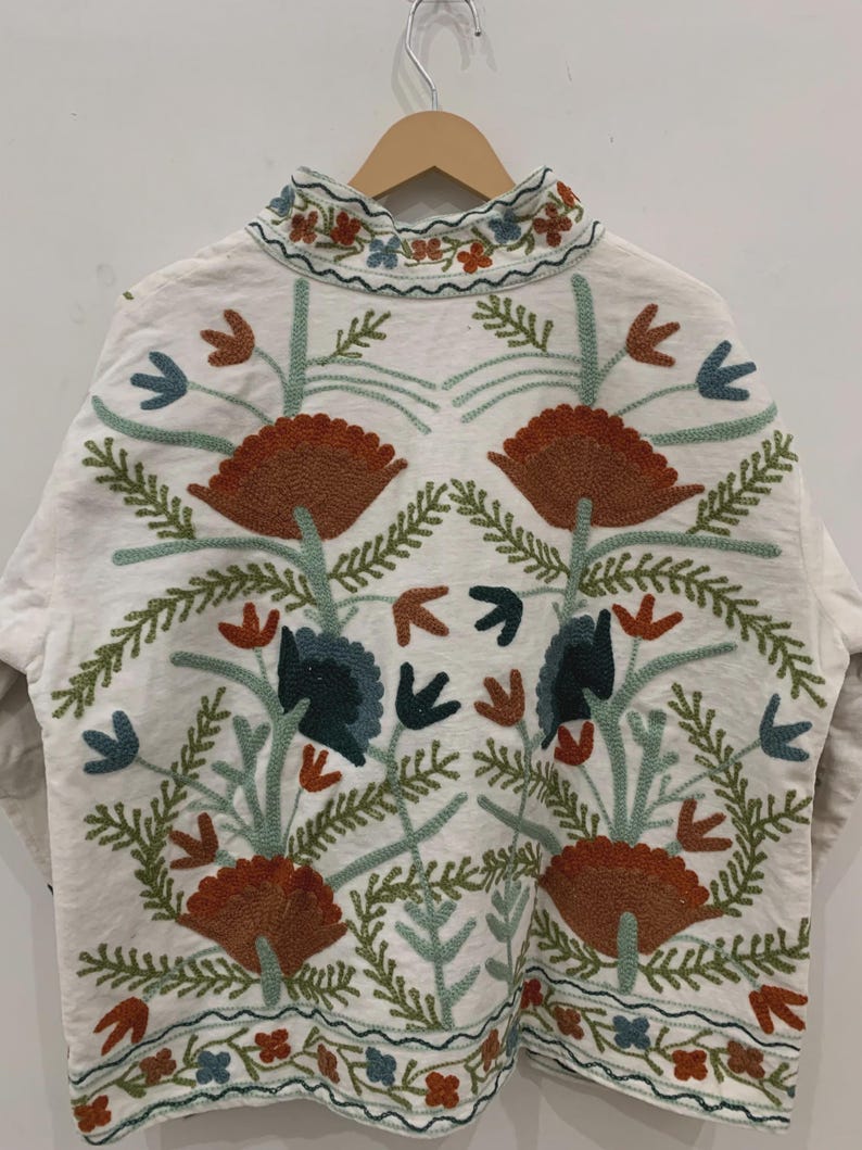 Puede incluir: Chaqueta blanca con intrincados bordados florales en tonos naranja, azul y verde. La chaqueta presenta un cuello alto y patrones detallados, mostrando un estilo bohemio o de arte popular. El bordado incluye flores y hojas estilizadas.