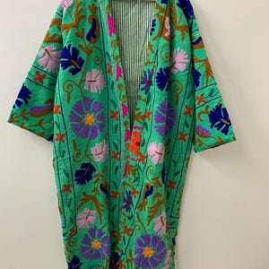 May include: A long, open-front robe in a vibrant green hue, adorned with intricate floral embroidery in shades of purple, pink, blue, and brown. The robe features a striped lining and is displayed on a wooden hanger, showcasing its detailed craftsmanship.
