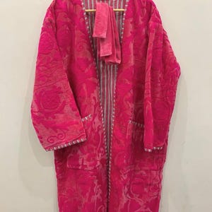 May include: A bright pink robe with a matching sash, displayed on a wooden hanger. The robe has an embossed floral pattern and two front pockets. The interior and trim feature a striped design.