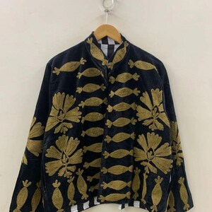 May include: Black jacket with gold floral and fish-shaped embroidery. The jacket has a high collar and a zipper closure. The lining is black and white checkered. The jacket is hanging on a wooden hanger.